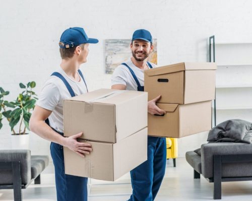 two-movers-in-uniform-looking-at-each-other-while-carrying-cardboard-boxes-in-apartment.jpg
