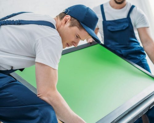 two-movers-in-uniform-transporting-tv-with-green-screen-in-apartment.jpg